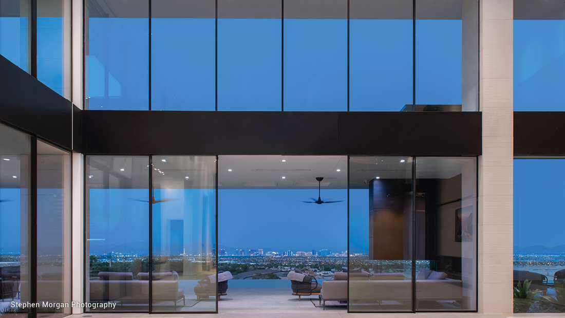 Luxury modern home with floor-to-ceiling glass walls offering panoramic city views at dusk, featuring sleek minimalist interior and outdoor seating area.