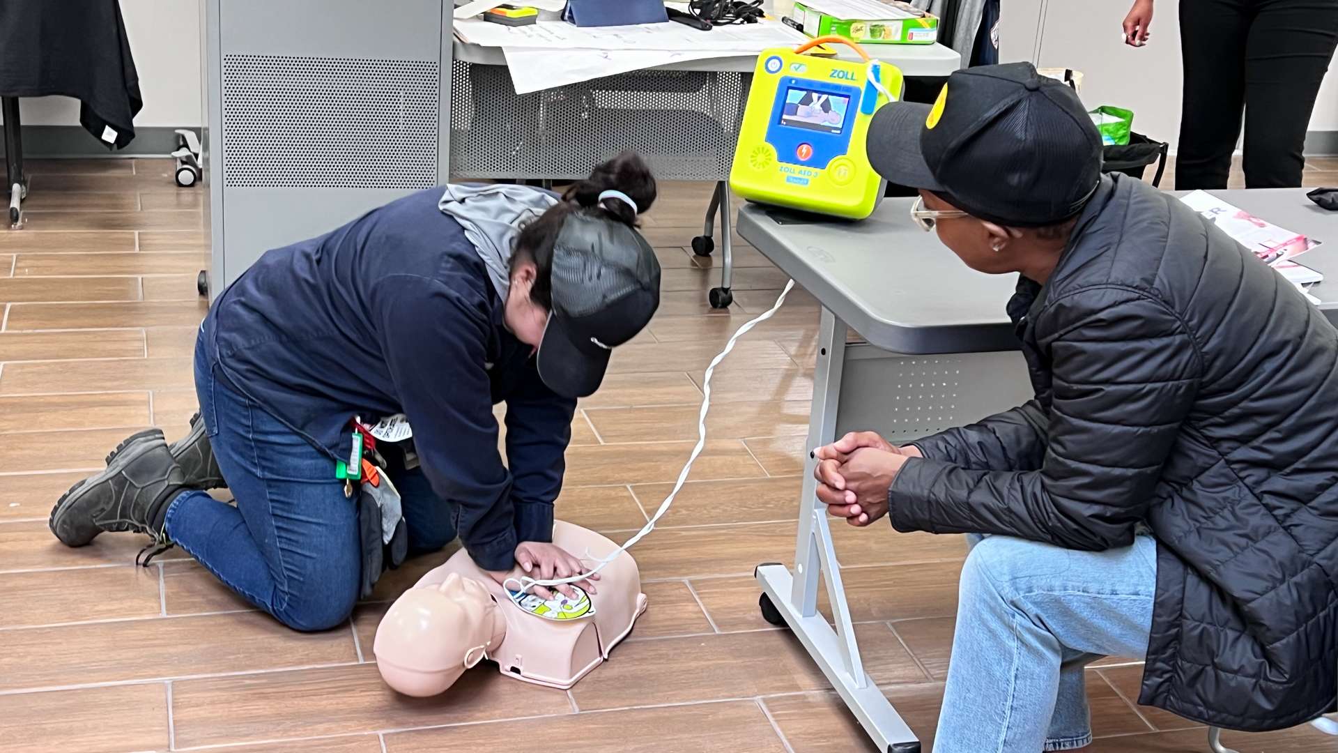 Person kneeling on a wood-look floor performing chest compressions on a CPR manikin during a training session, with an AED device on a nearby table and another participant observing.