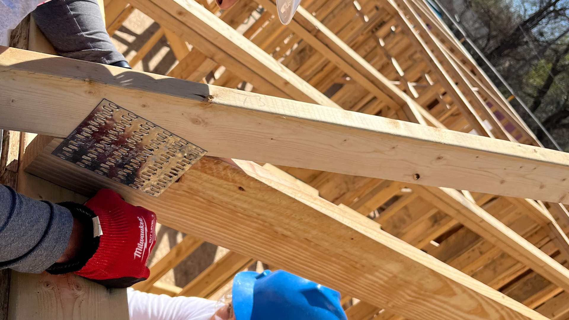 Volunteer wearing a white hard hat and red gloves securing a metal connector plate on wooden roof trusses at a building site, with another volunteer in a blue hard hat visible below.