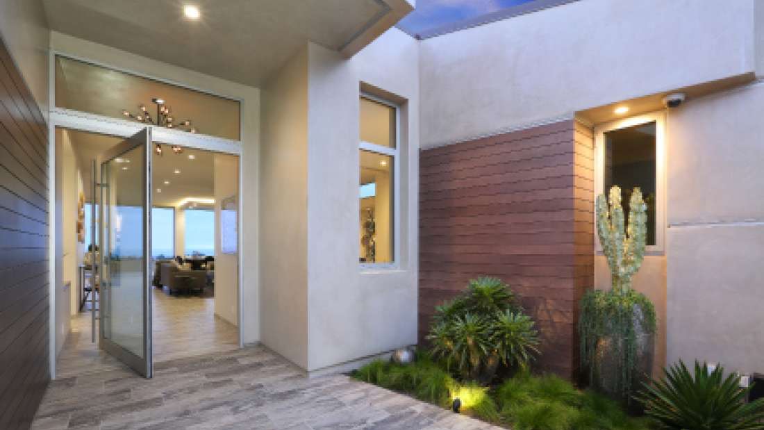 Modern home entryway with pivot glass door, beige stucco walls, horizontal wood siding accent, and minimalist landscaping featuring desert plants and recessed lighting.