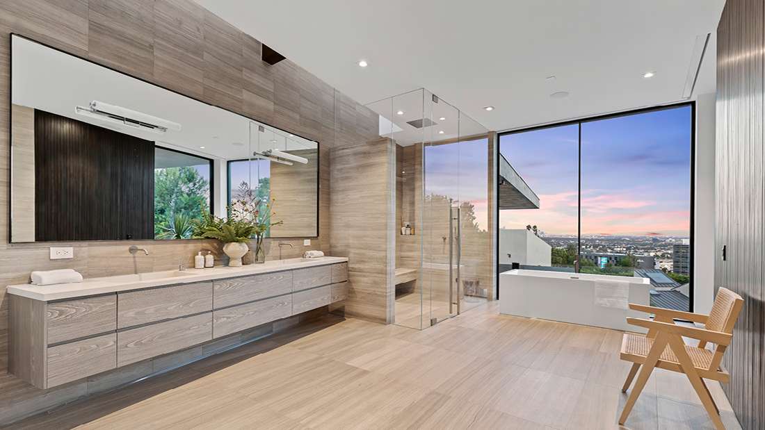 Luxury bathroom with double vanity, large mirror, glass shower enclosure, freestanding bathtub, and floor-to-ceiling windows offering panoramic city views at sunset.