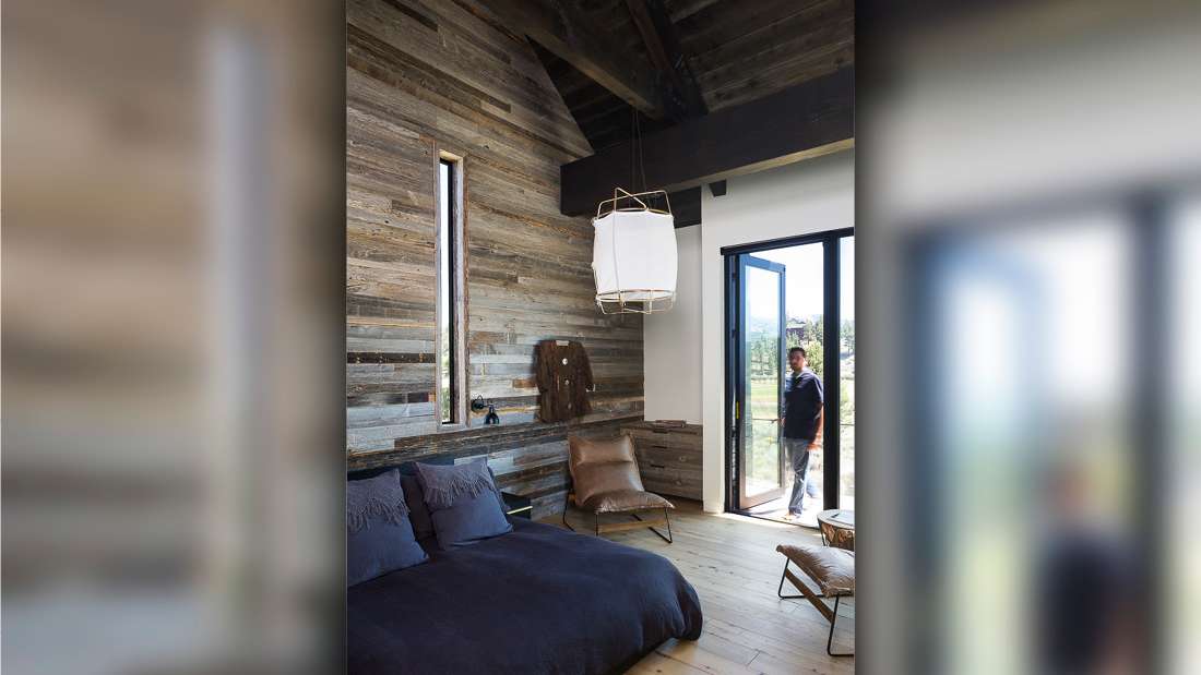 Rustic modern bedroom with reclaimed wood accent wall, vaulted ceiling with exposed beams, dark bedding, leather lounge chair, and folding glass doors opening to outdoor balcony with scenic views.