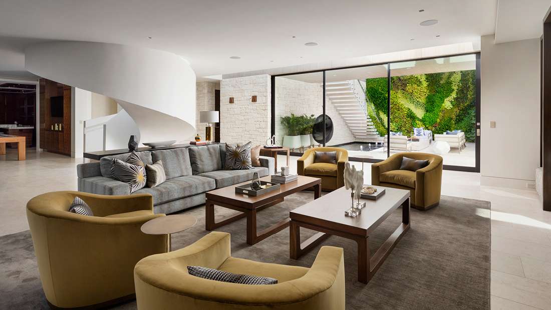 Luxury modern living room with curved white staircase, gray sectional sofa, mustard armchairs, wooden coffee tables, and floor-to-ceiling glass doors opening to a courtyard with vertical garden.