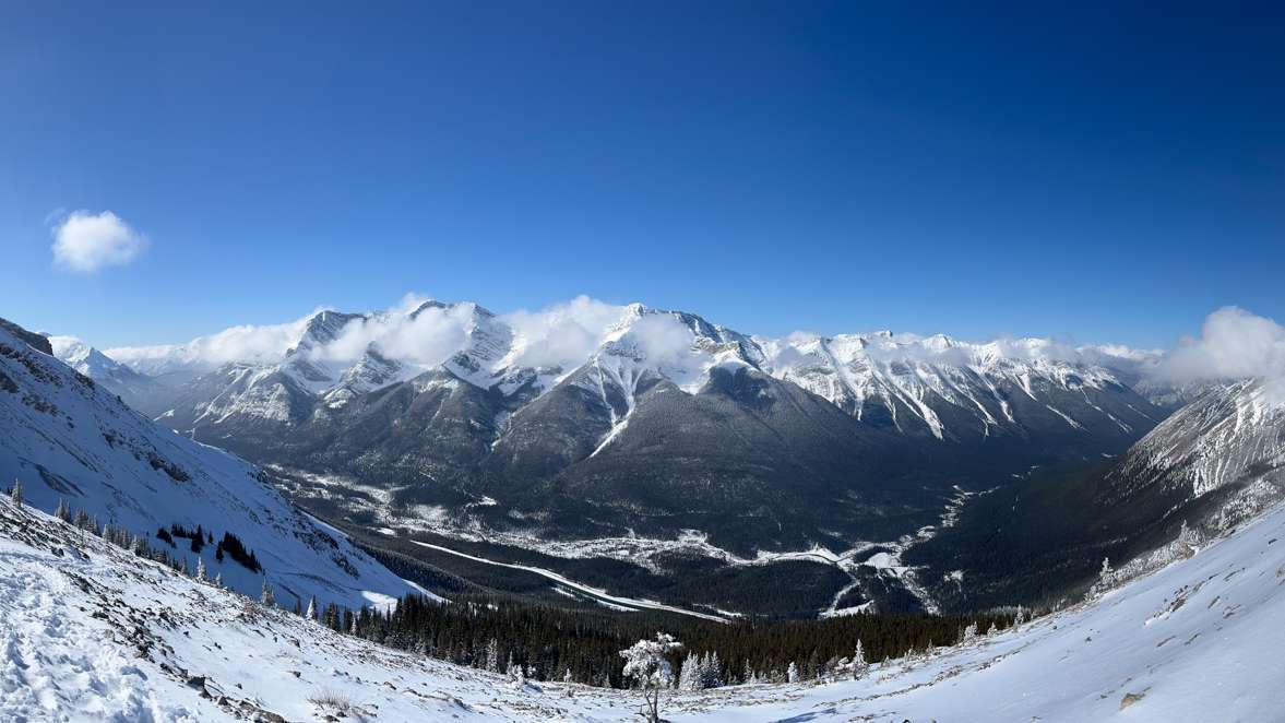 Panoramic view of snow-covered mountain slopes and a valley with winding trails under a clear blue sky and scattered clouds.  