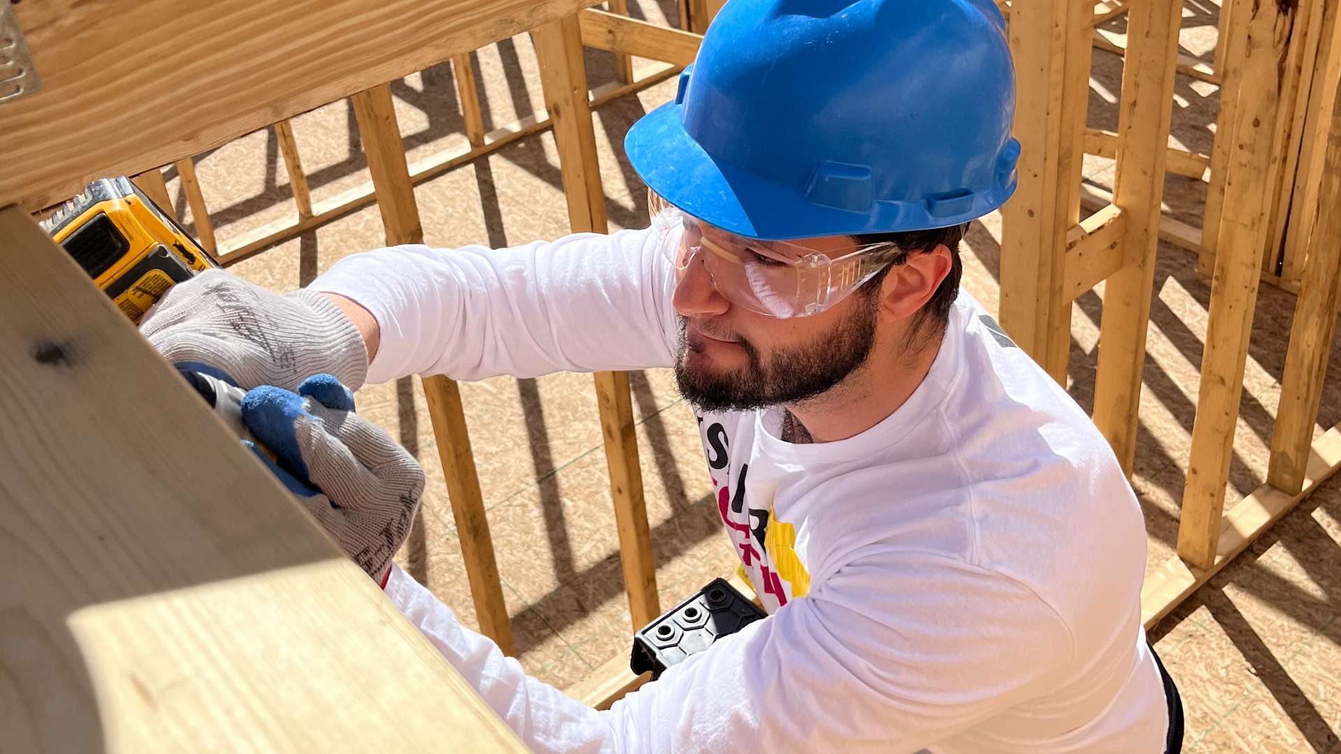 Volunteer wearing a blue hard hat and gray gloves using a power drill to secure wooden framing at a building site with exposed lumber structure.