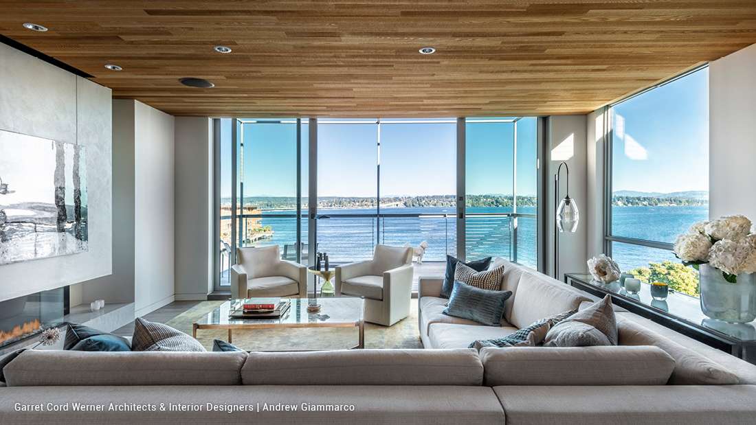 Luxury waterfront living room with floor-to-ceiling glass walls, panoramic ocean views, light gray sectional sofa, marble coffee table, and wood-paneled ceiling.