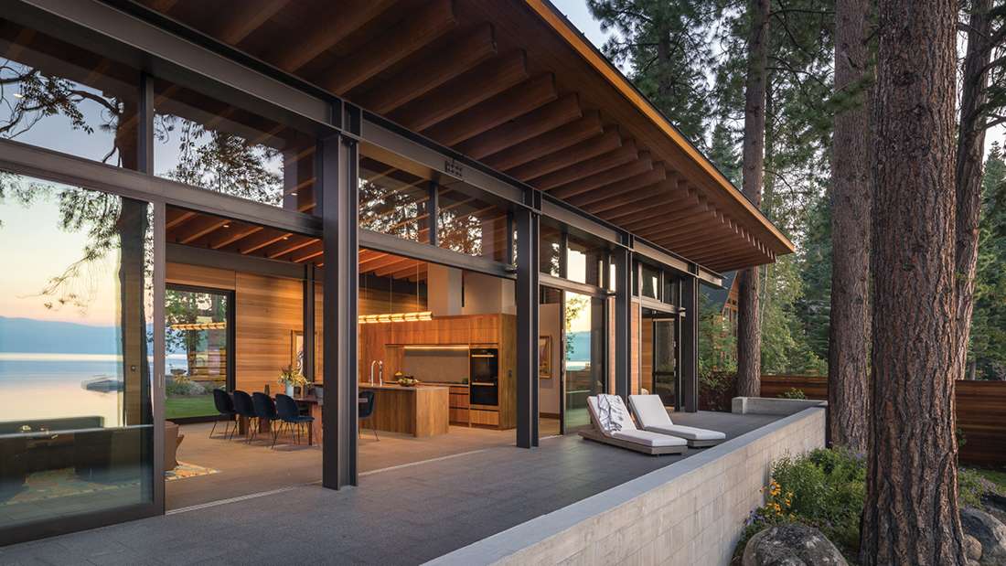 Modern lakefront home with exposed wood ceiling beams, floor-to-ceiling glass walls, open-concept kitchen with wood cabinetry, and outdoor patio featuring lounge chairs overlooking water and forest views.