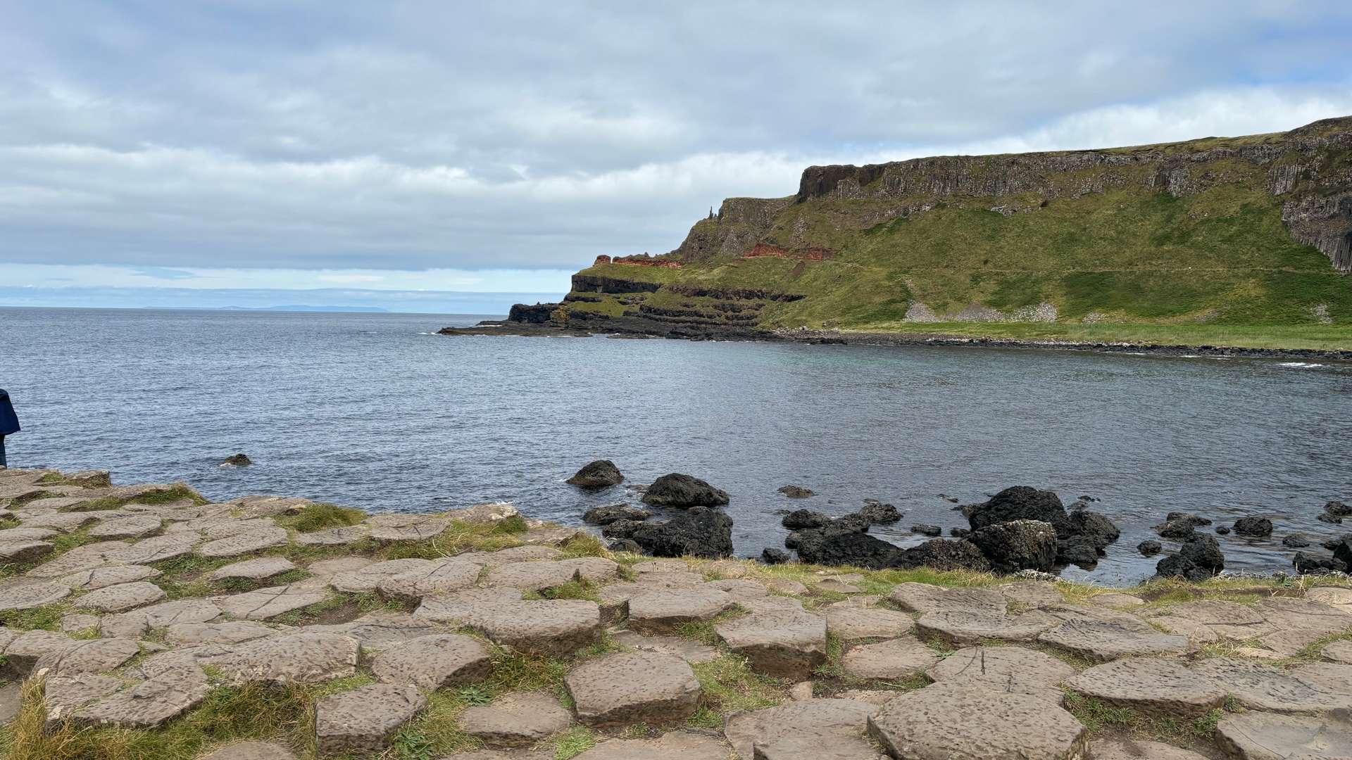 Coastal view with hexagonal stone formations in foreground, calm sea, and grassy cliffs under a cloudy sky.