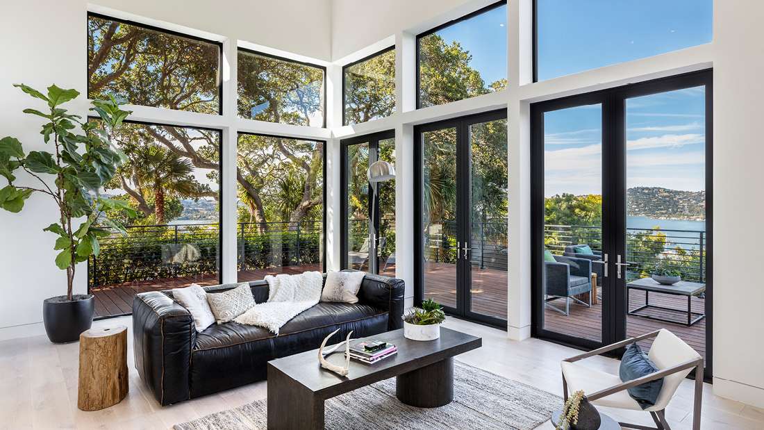 Modern living room with floor-to-ceiling glass doors and windows offering panoramic views of trees and water, black leather sofa with white throw, minimalist coffee table, and outdoor deck with lounge seating.