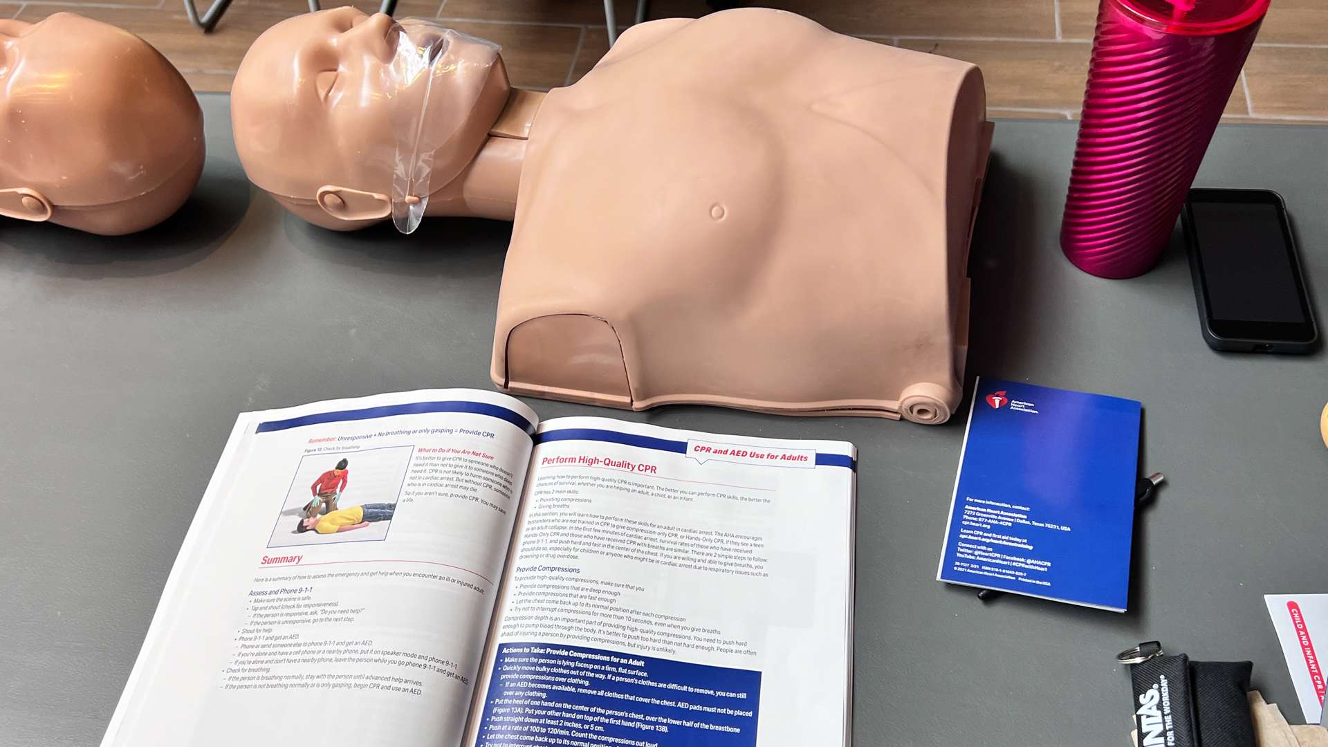 CPR training setup with a torso manikin, an open CPR instruction manual, a pink tumbler, a smartphone, and a blue booklet on a gray table.
