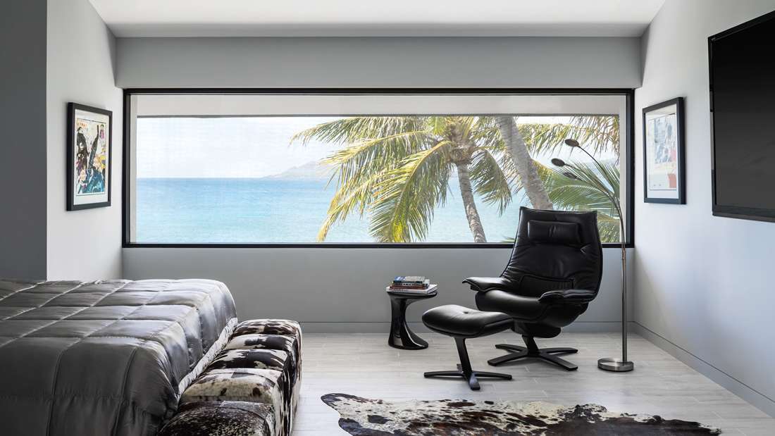 Modern coastal bedroom with wide horizontal window framing ocean and palm tree views, gray bedding, black leather lounge chair with matching ottoman, cowhide rug, and minimalist decor.