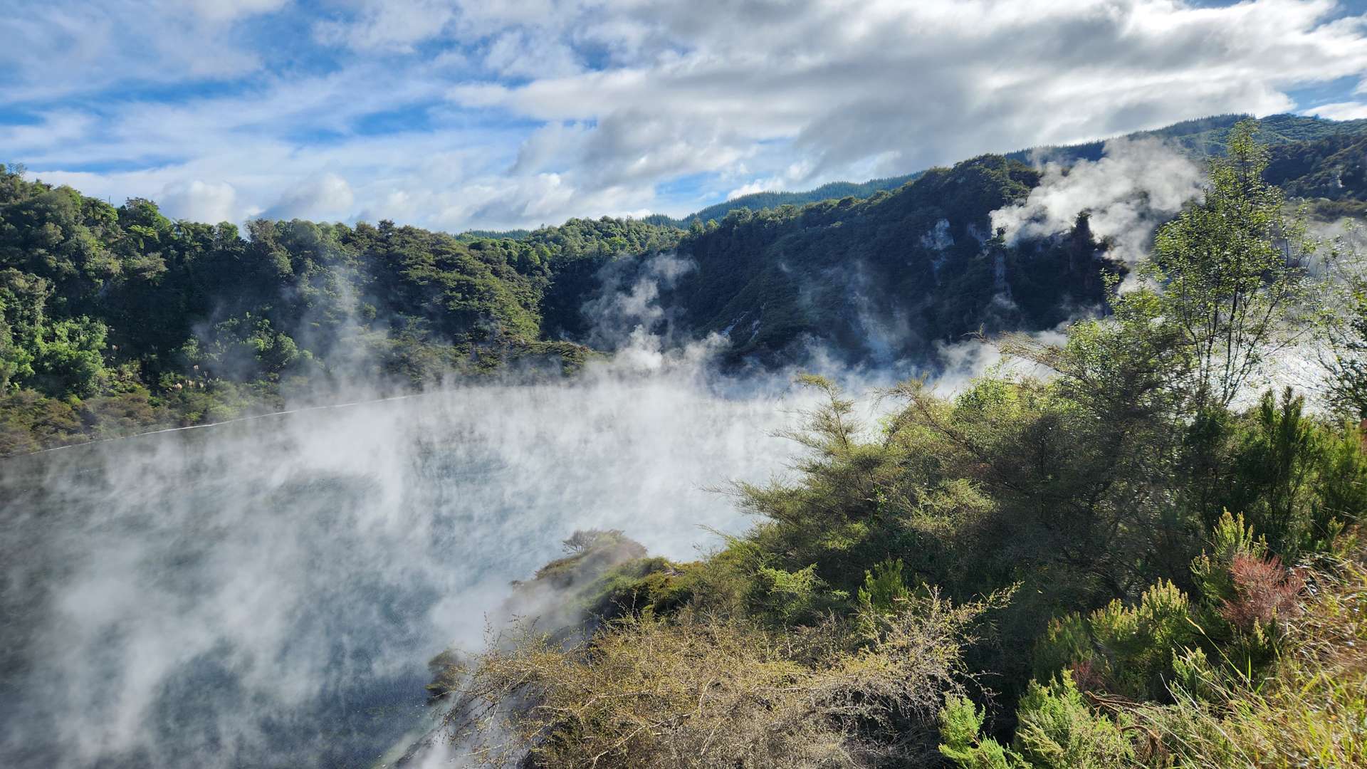 Geothermal lake with rising steam surrounded by lush green hills under a bright blue sky with scattered clouds