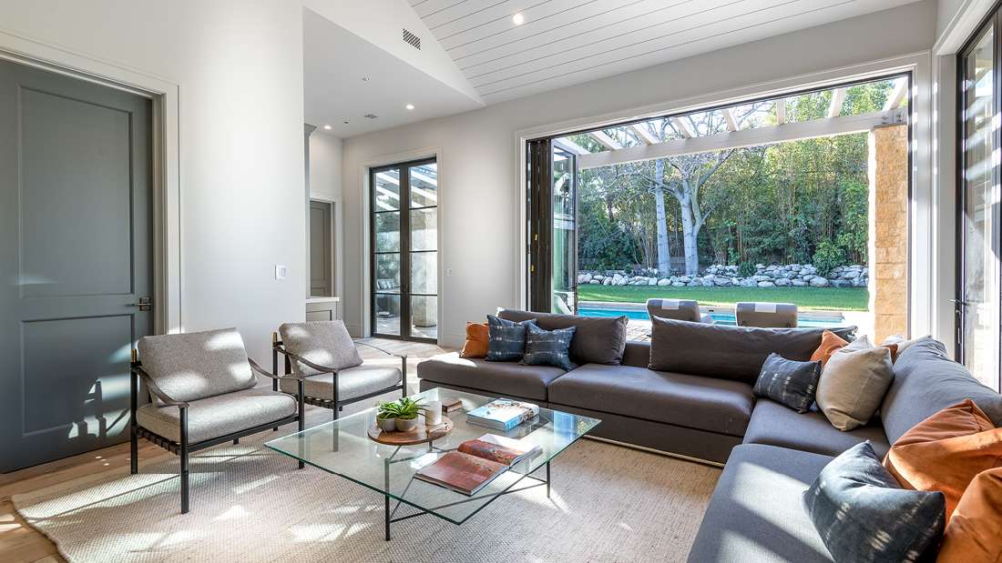 Modern living room with large gray sectional sofa, glass coffee table, and two upholstered armchairs, featuring folding glass doors opening to a backyard with pool and lush greenery.