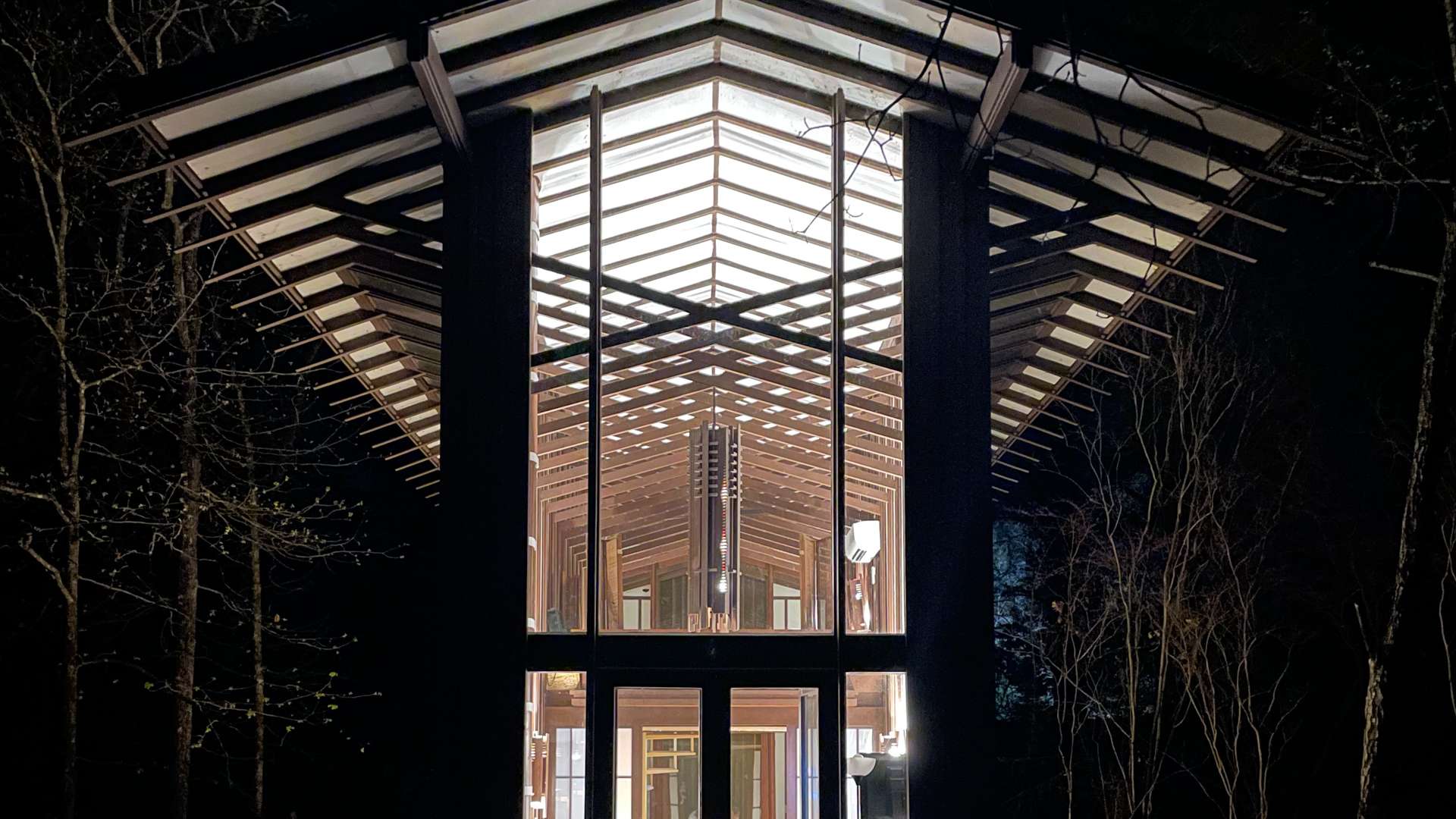   Modern glass pavilion illuminated at night, showing people seated inside under a striking geometric roof structure.