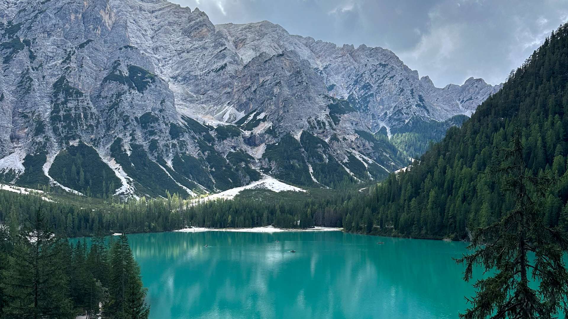 Turquoise mountain lake surrounded by dense pine forest and towering rocky peaks under a partly cloudy sky.