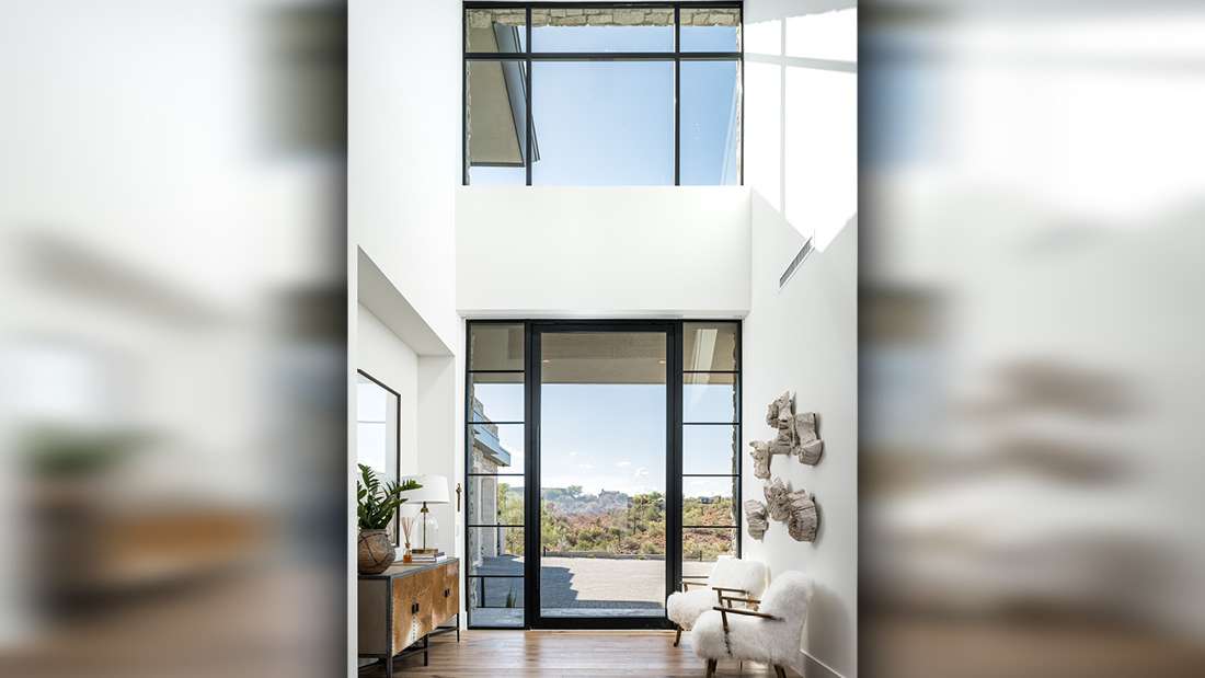Modern entryway with double-height ceiling, large black-framed glass doors and windows, white walls, wooden console table with decorative accents, and cozy armchair with textured throw overlooking scenic hillside views.