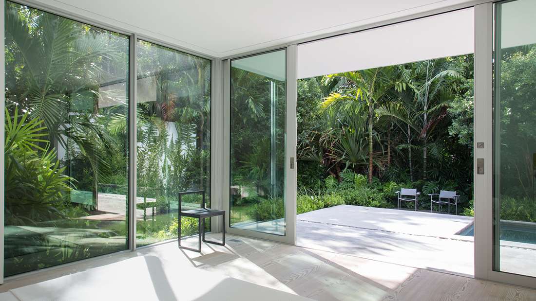 Modern indoor-outdoor living space with large sliding glass doors opening to a tropical garden and pool area, featuring minimalist white walls, light wood flooring, and a single black chair.