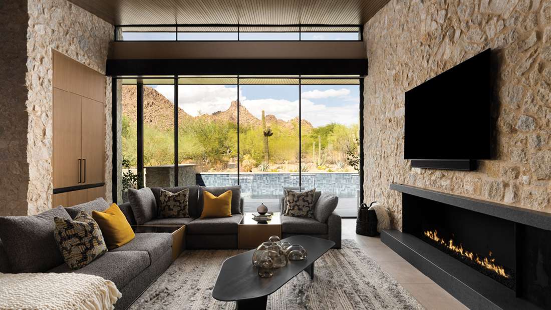 Modern living room with stone accent walls, large windows overlooking desert mountains, sleek fireplace, and contemporary gray sectional sofa.