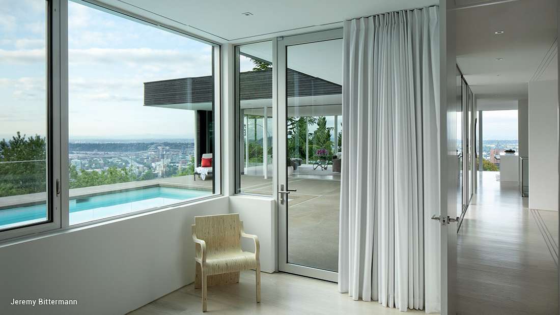 Modern interior with floor-to-ceiling glass doors and windows offering panoramic city views, white curtains, light wood flooring, and outdoor pool visible from the living space.