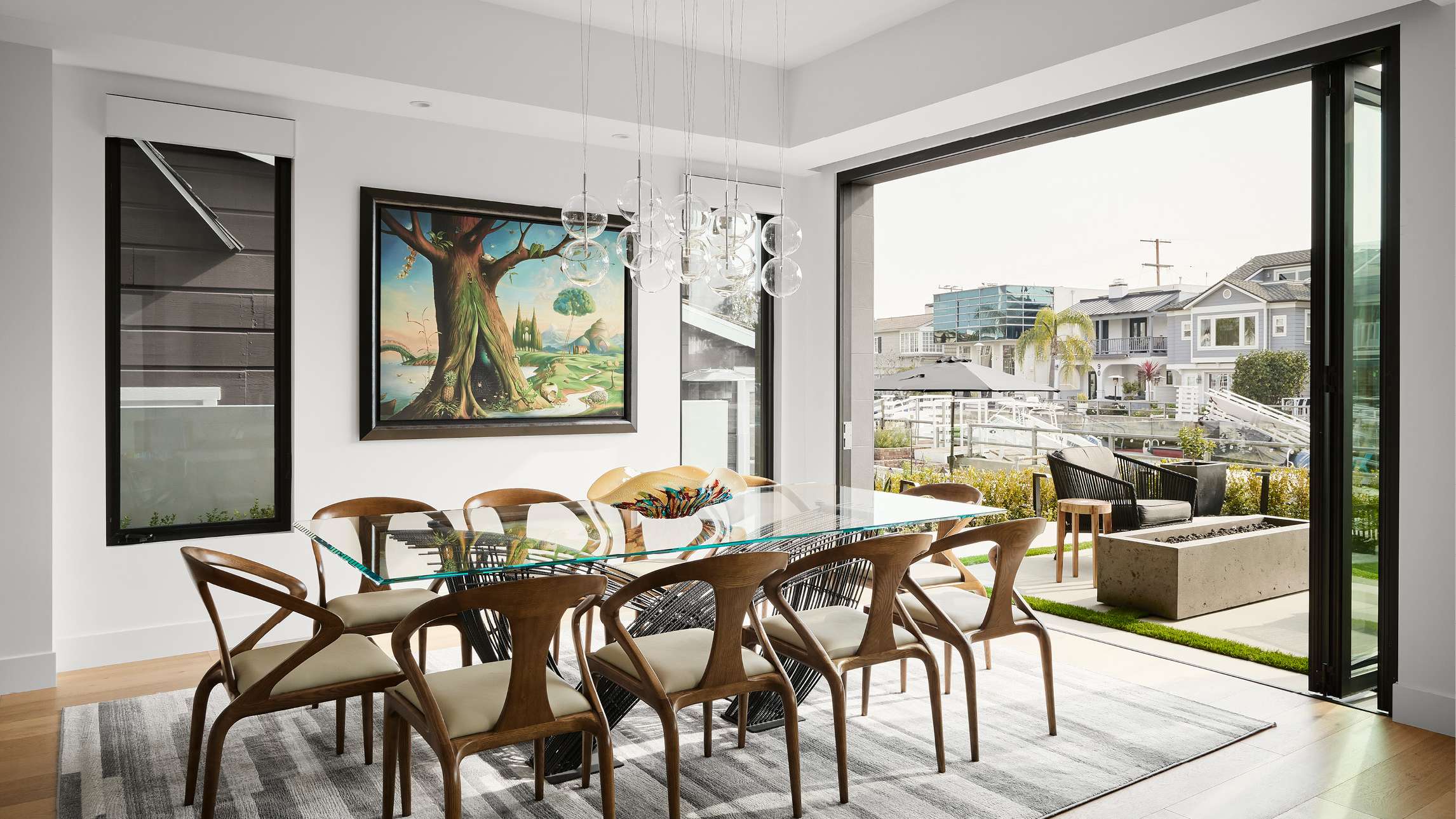 Modern dining room with glass-top table seating eight wooden chairs, pendant chandelier with glass globes, large folding glass doors opening to outdoor patio with fire pit and lounge chairs overlooking waterfront homes.