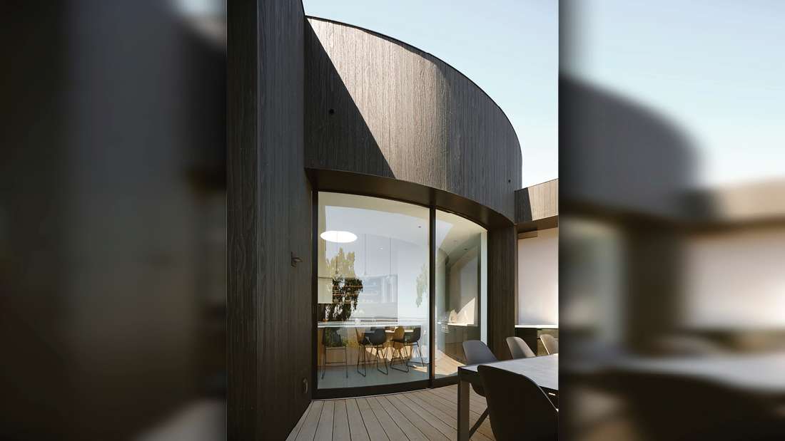 Contemporary home exterior with curved architectural design, dark wood cladding, and large floor-to-ceiling glass doors opening to a deck with outdoor dining area.