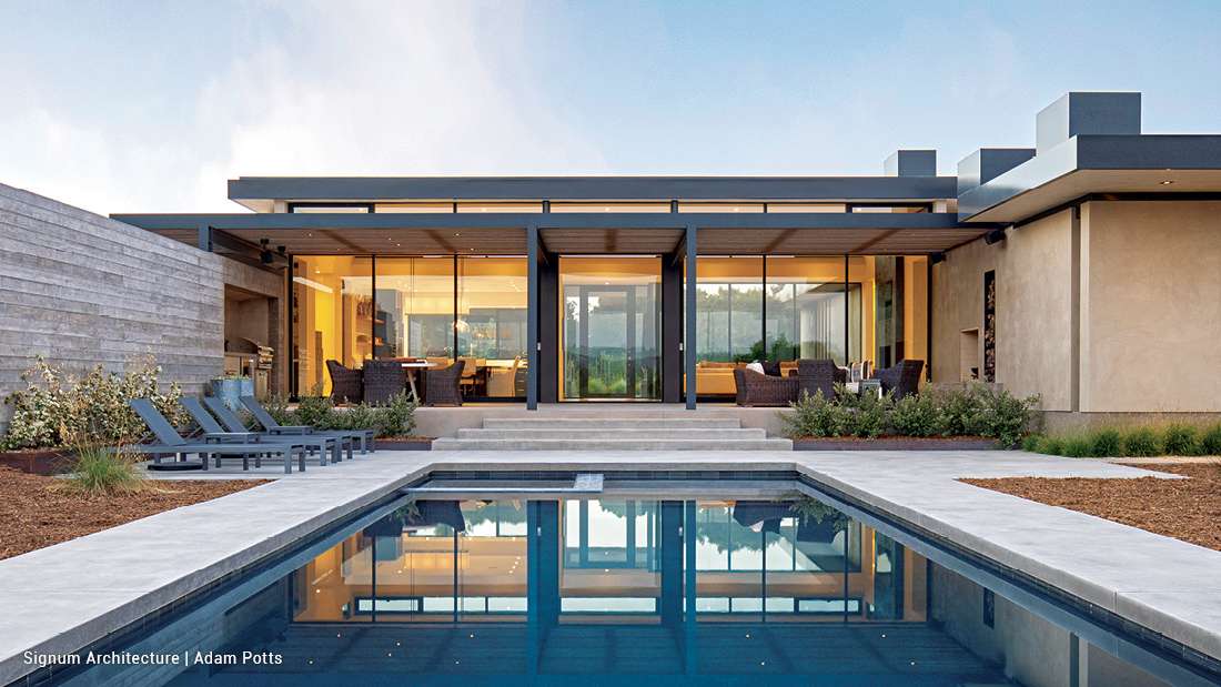 Modern single-story luxury home with floor-to-ceiling glass walls, flat roof design, outdoor lounge seating, and rectangular swimming pool reflecting warm interior lighting.