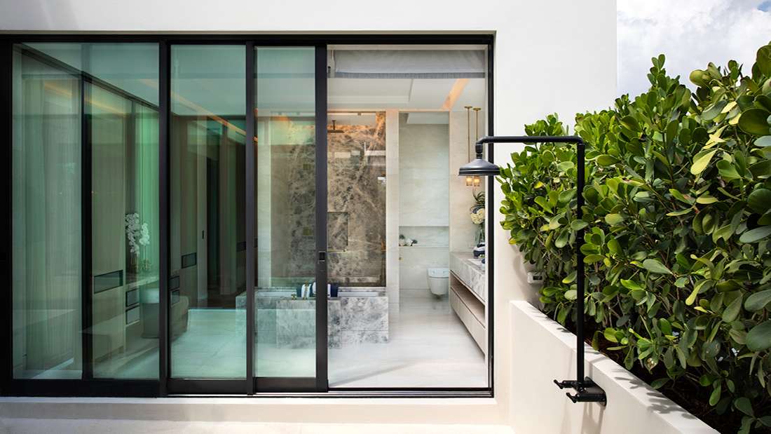 Luxury bathroom with floor-to-ceiling sliding glass doors, marble shower enclosure, floating vanity with double sinks, and outdoor black rain shower surrounded by lush green plants.