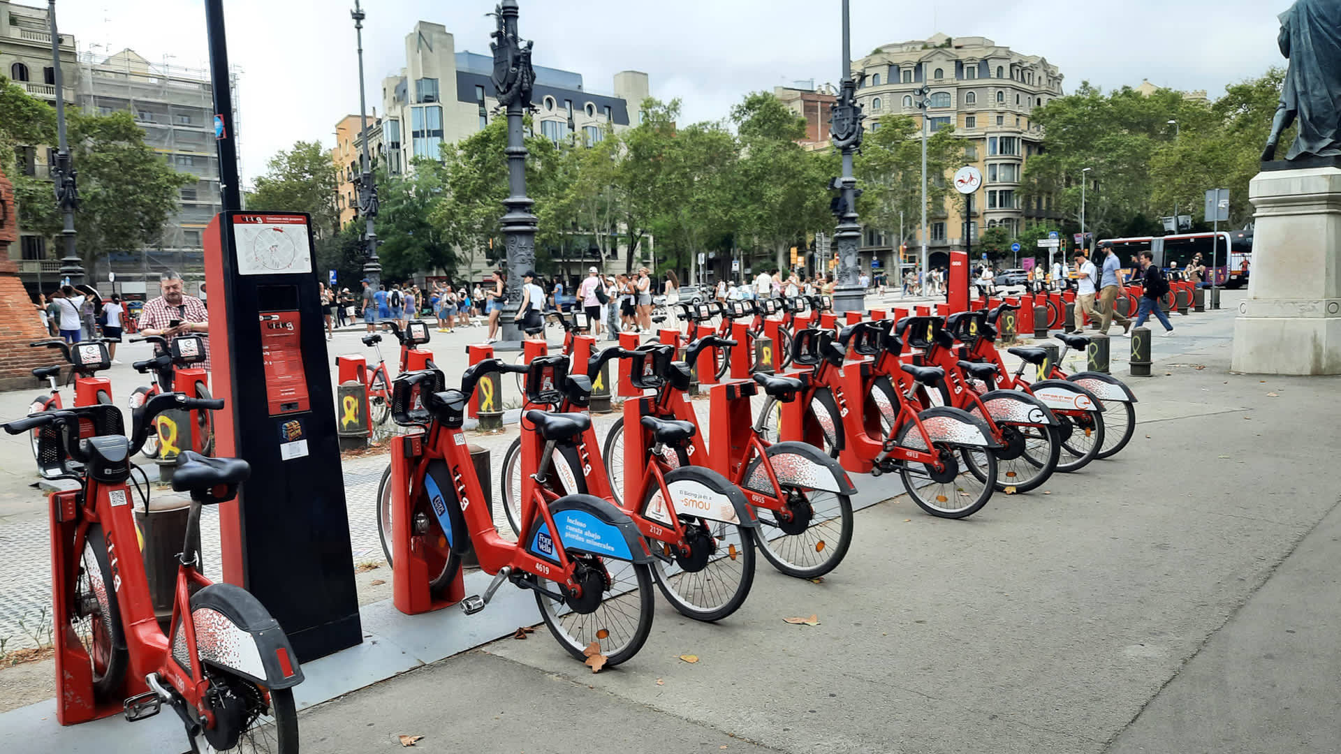 Row of red and black rental bicycles parked at a bike-sharing station in a busy urban square, with people walking in the background, trees lining the street, and historic buildings visible under a cloudy sky.