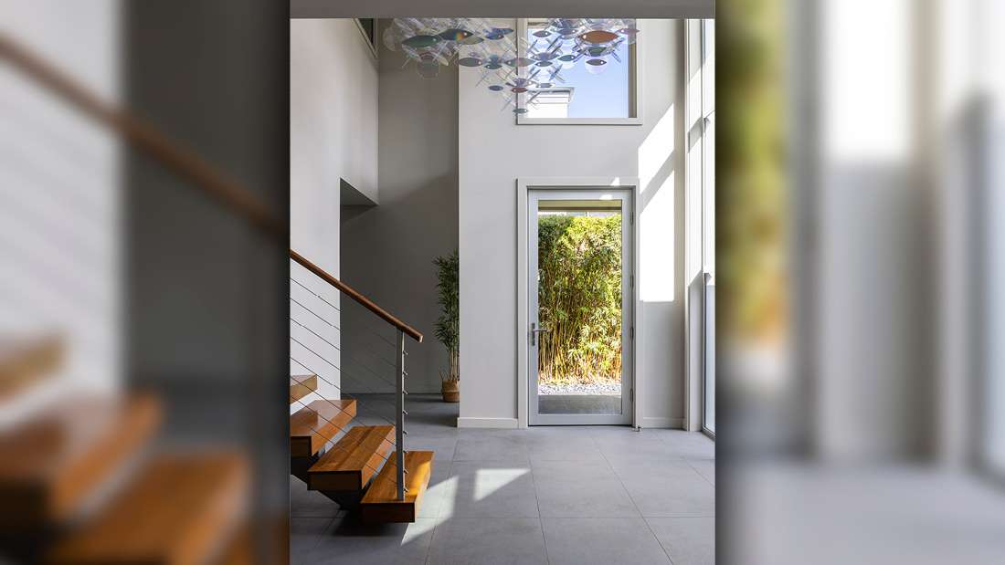 Modern entryway with floor-to-ceiling glass door, minimalist white walls, large gray tile flooring, and floating wooden staircase with metal railing under natural light.