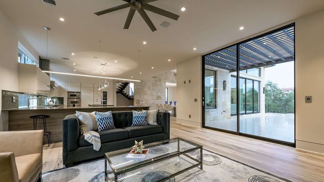 Elegant open-concept living area with modern kitchen, glass sliding doors leading to a covered patio, and stylish gray sofa with patterned cushions.