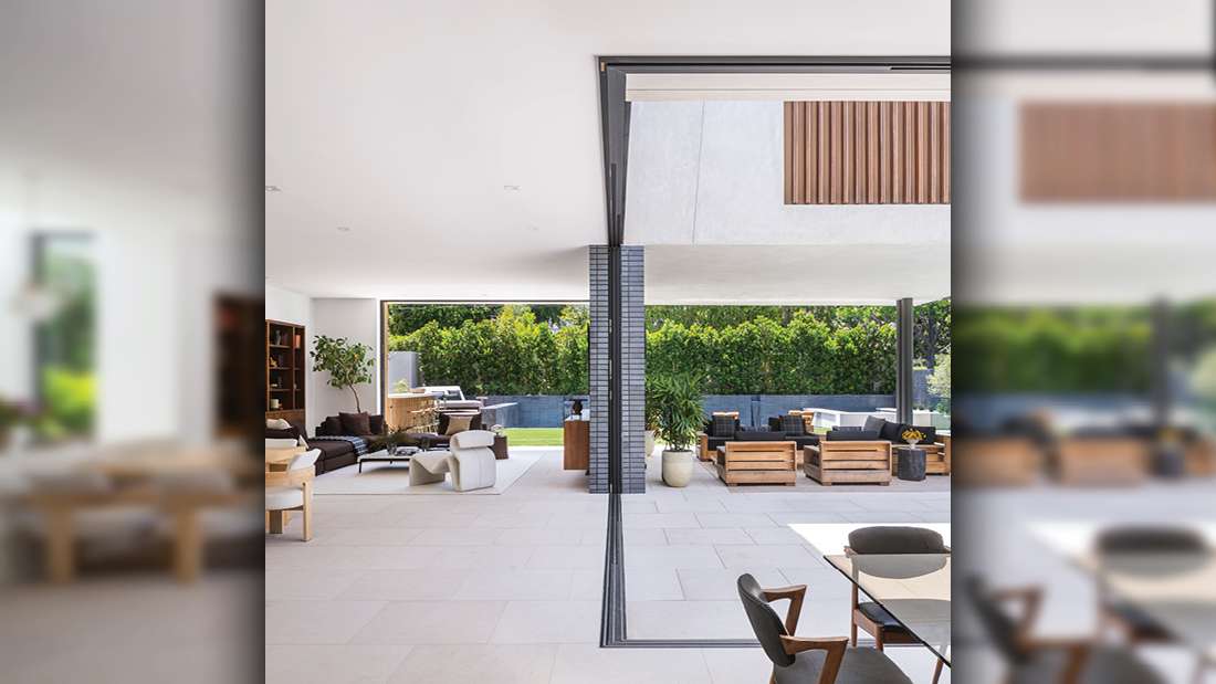 Luxury open-concept indoor-outdoor living space with sliding glass walls, modern lounge furniture, dining area with glass table, and landscaped greenery in the background.