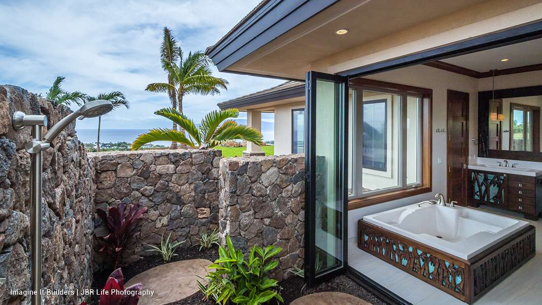 Luxury outdoor bathroom with stone privacy walls, stainless steel rain shower, tropical landscaping, and indoor soaking tub framed by folding glass doors overlooking ocean views.