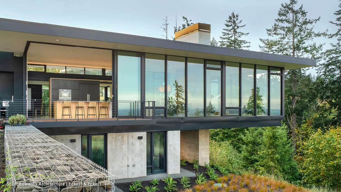 Contemporary hillside home with cantilevered design, floor-to-ceiling glass walls, flat roof, and open kitchen with wood island and bar stools overlooking forest views.