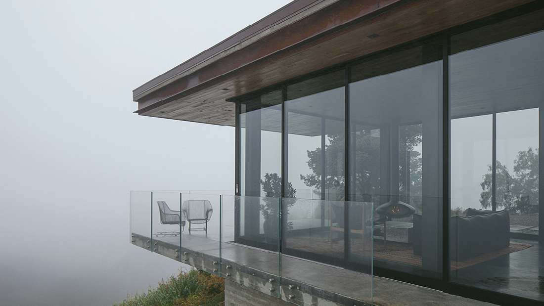 Modern cliffside home with cantilevered concrete balcony, glass railing, and floor-to-ceiling glass walls overlooking foggy ocean views.