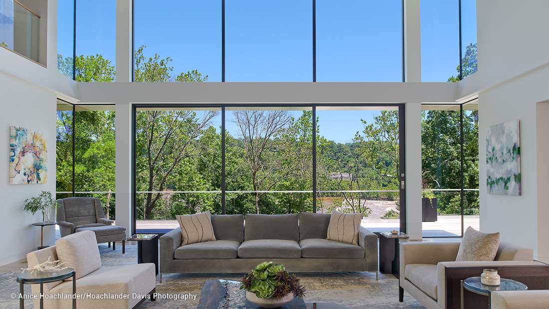 Luxury double-height living room with floor-to-ceiling glass walls, panoramic forest views, modern gray sofa set, neutral armchairs, and abstract wall art.