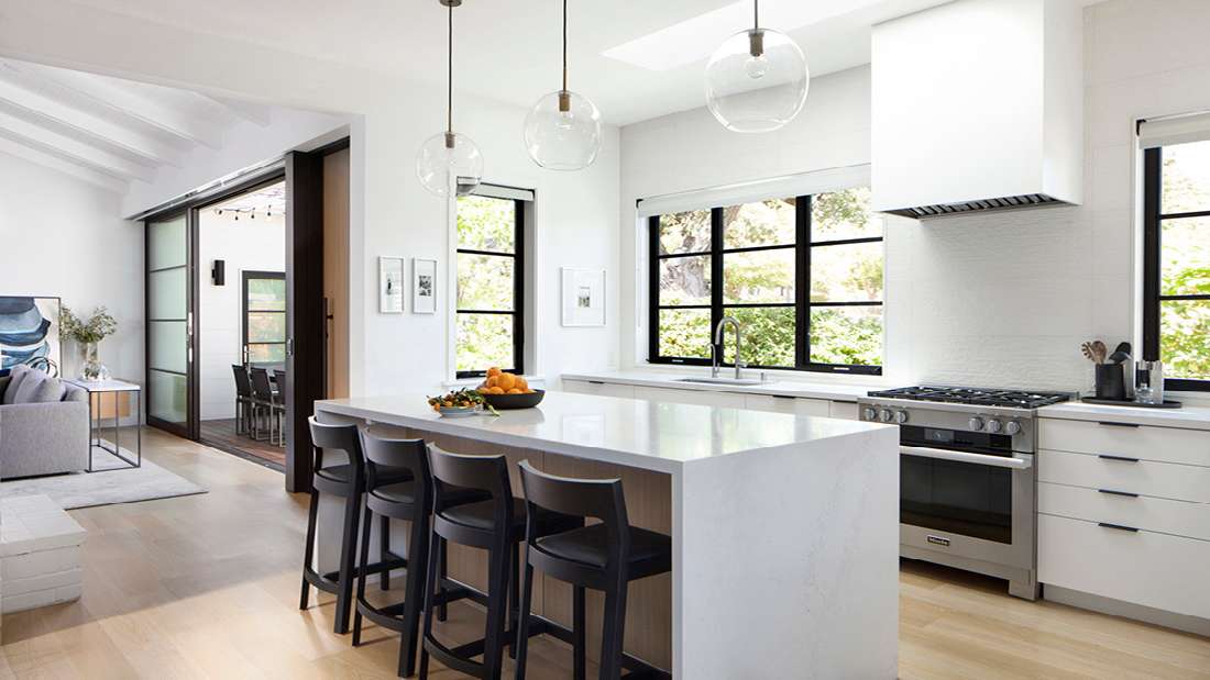 Modern kitchen with large white waterfall island, four black barstools, pendant glass globe lights, black-framed windows, and stainless steel gas range in an open-concept layout.