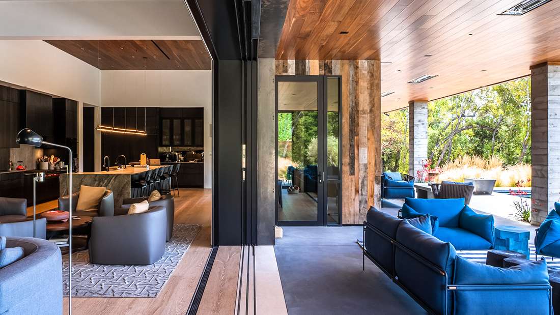 Luxury open-concept living and kitchen area with natural wood ceiling, black cabinetry, marble island with barstools, and folding glass doors opening to a covered patio featuring blue lounge chairs and outdoor greenery.