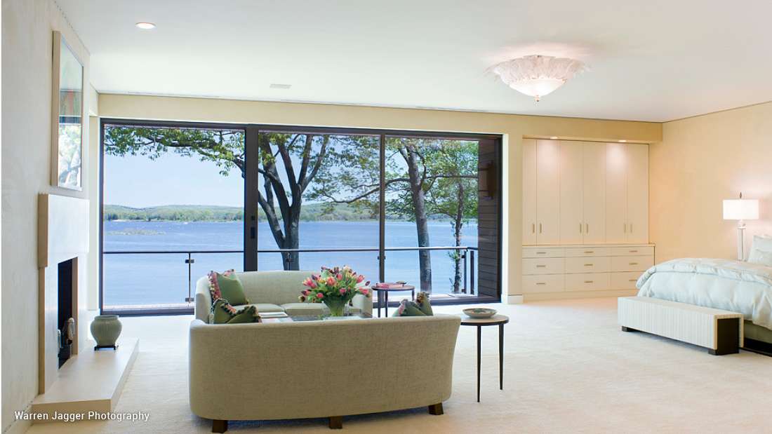 Spacious lakefront bedroom with sitting area, large sliding glass doors opening to water views, built-in cabinetry, and soft neutral decor.