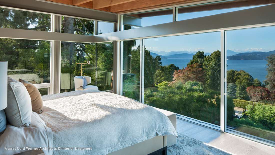 Luxury bedroom with floor-to-ceiling glass walls offering panoramic ocean and mountain views, exposed wood ceiling beams, white bedding, and minimalist decor.