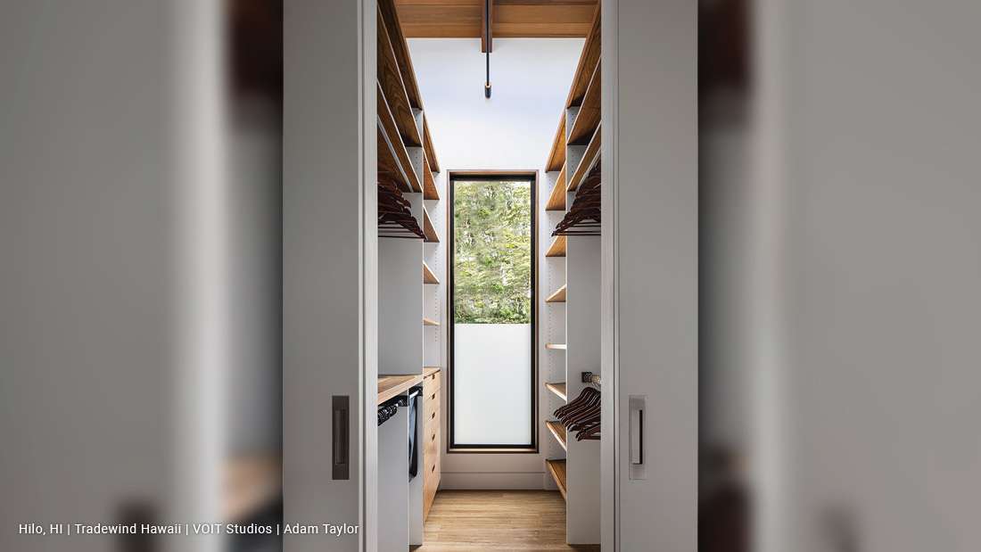 Modern walk-in closet with symmetrical wooden shelving, built-in drawers, and a tall narrow window featuring frosted glass on the lower half and clear glass above, providing natural light and greenery views.