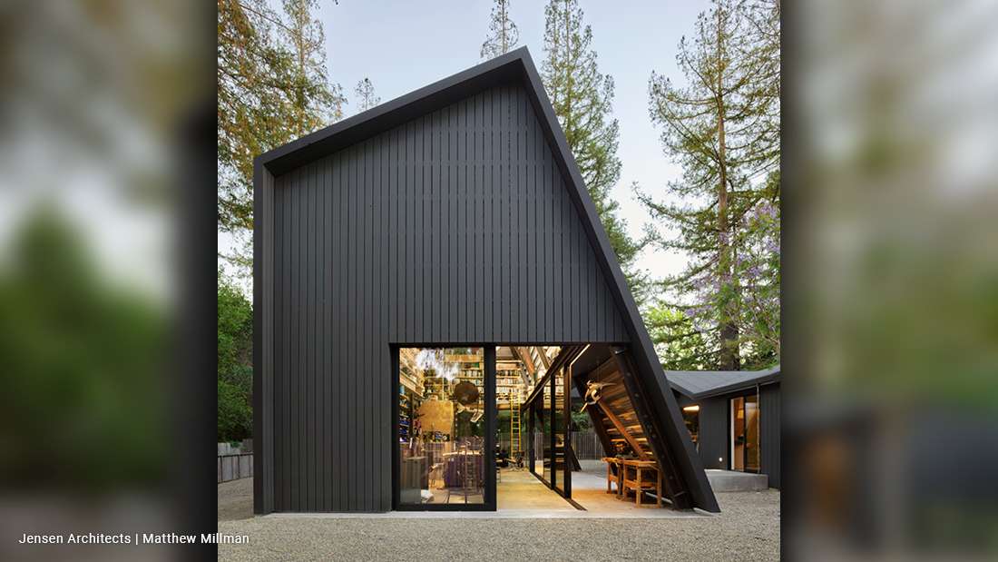 Modern A-frame style building with dark vertical wood siding, large glass windows, and exposed interior shelving, set among tall pine trees.