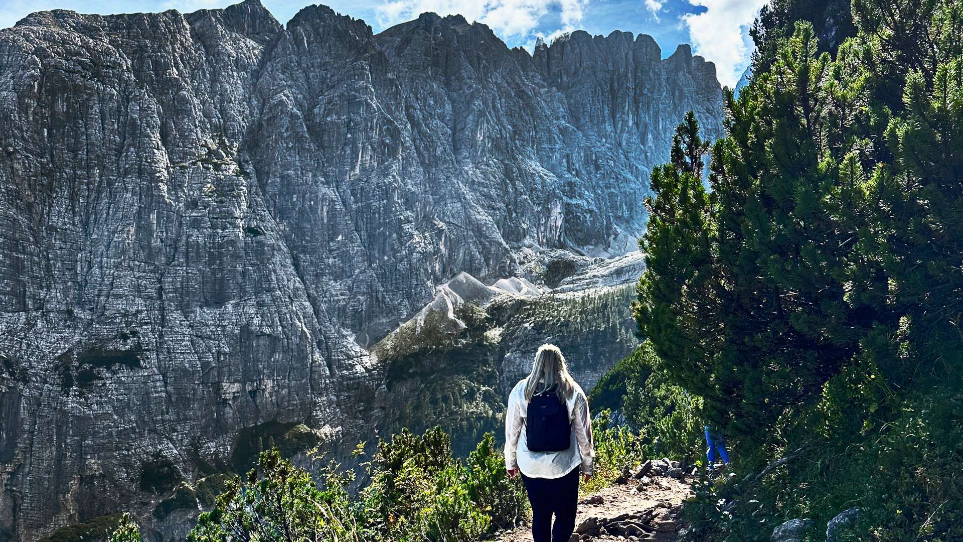 Person hiking along a rocky mountain trail surrounded by green shrubs with towering gray cliffs under a bright blue sky.