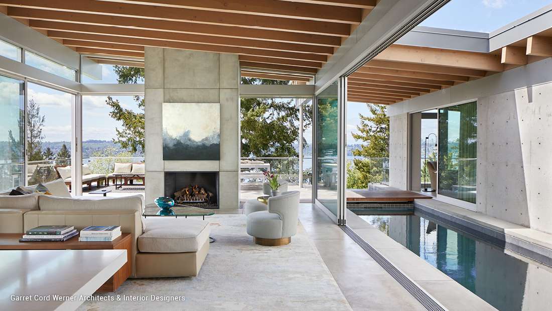 Luxury open-concept living space with exposed wood ceiling beams, concrete fireplace wall featuring abstract art, floor-to-ceiling glass doors, and indoor lap pool overlooking scenic lake views.