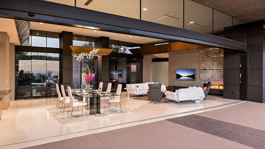 Luxury open-concept dining and living area with glass walls, modern chandelier, white leather sofas, and sleek fireplace overlooking panoramic sunset views.