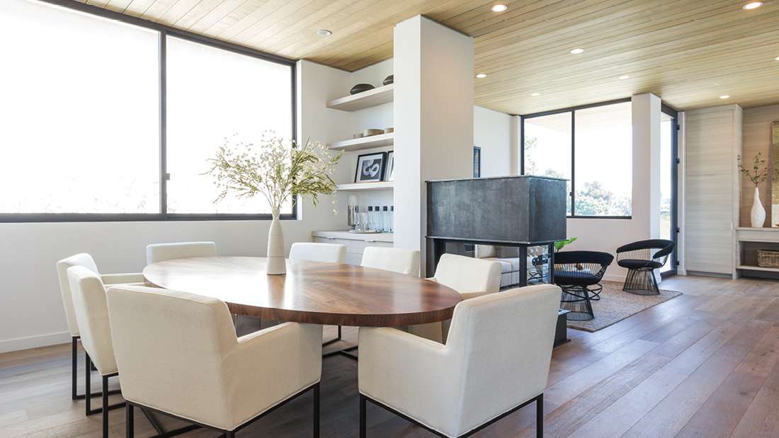 Modern dining area with round wooden table, six upholstered white chairs, light wood ceiling with recessed lighting, large windows with black frames, and open shelving displaying decor and glassware.