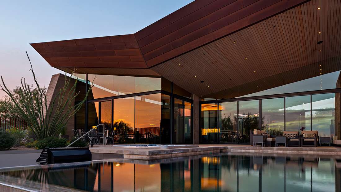 Contemporary desert home with angular roof design, floor-to-ceiling glass walls, outdoor seating area, and infinity pool reflecting a vibrant sunset.