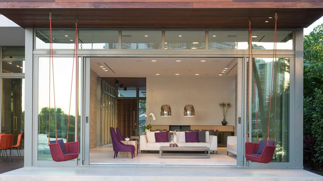Modern indoor-outdoor living space with large sliding glass doors, wood ceiling accents, and contemporary furniture including white sofa and purple chairs, featuring two red hanging swings and open view to lush greenery.