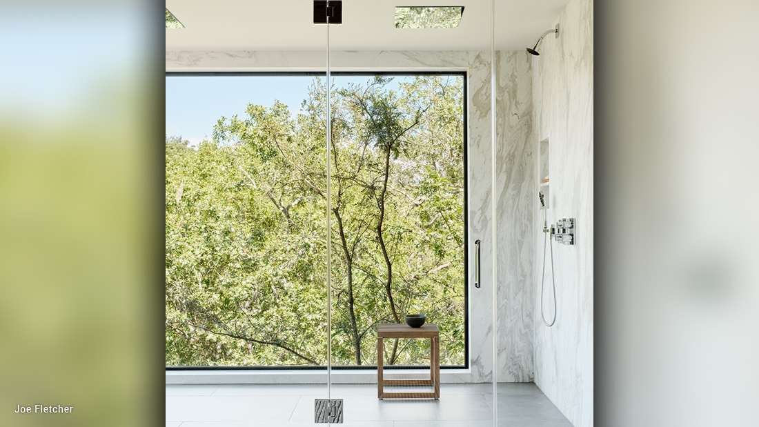 Luxury walk-in shower with floor-to-ceiling glass enclosure, large picture window offering lush forest views, marble walls, and minimalist wooden stool.
