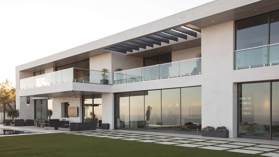 Modern two-story luxury home with minimalist white exterior, glass balcony railings, floor-to-ceiling windows, and outdoor lounge area next to a swimming pool.
