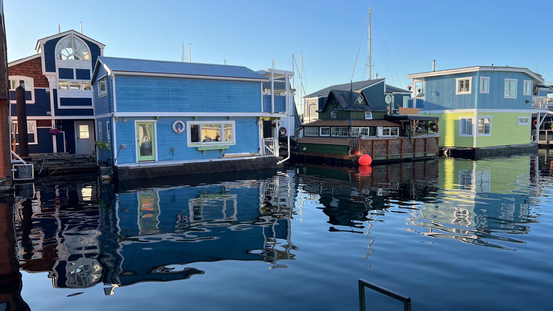 Colorful floating homes reflected on calm water in a marina under clear blue sky.